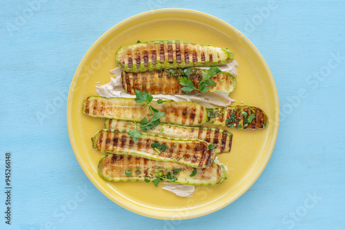 Grilled zucchini served with tahini sauce and chopped parsley on yellow platter over wooden background.