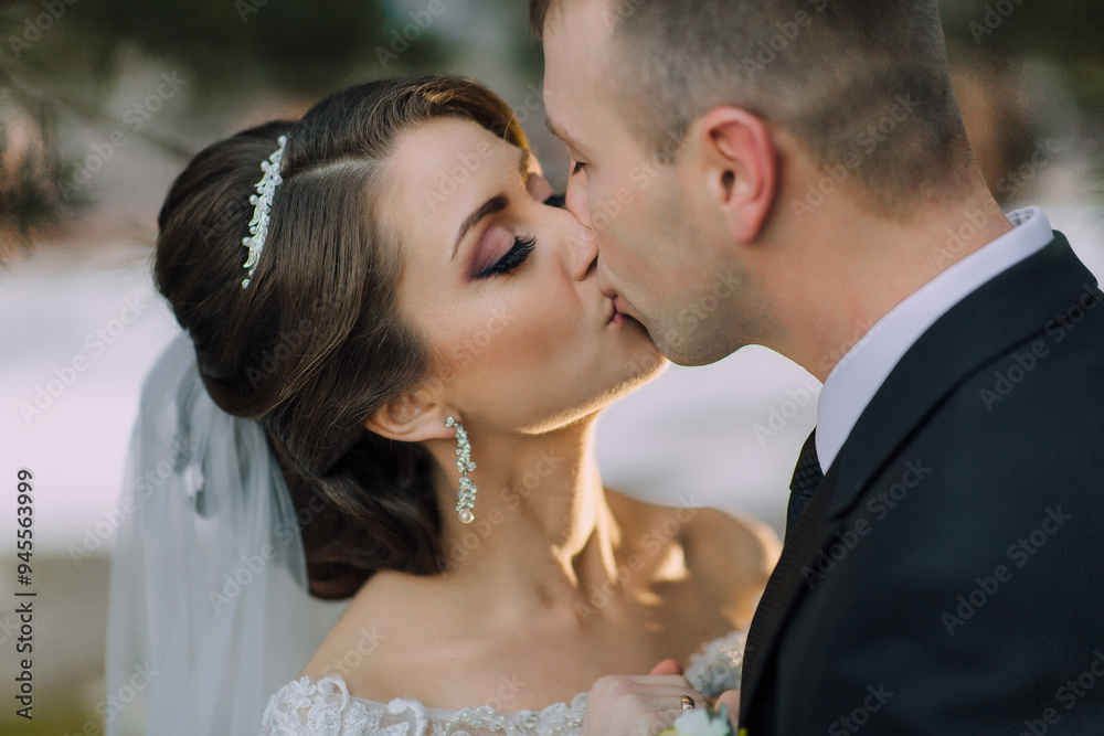 A bride and groom kiss in front of a wedding cake. The bride is wearing a white dress and the groom is wearing a black suit A bride and groom kiss in front of a wedding cake. The bride is wearing a white dress and the groom is wearing a black suit