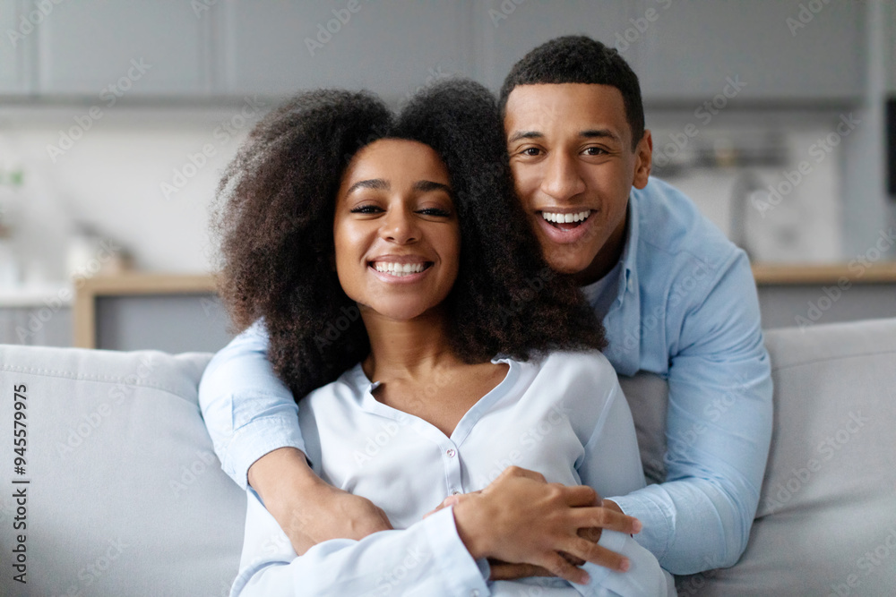 Happy black man embracing her wife and smiling to camera, family sitting on sofa, enjoying weekend together. Happiness in marriage Happy black man embracing her wife and smiling to camera, family sitting on sofa, enjoying weekend together. Happiness in marriage