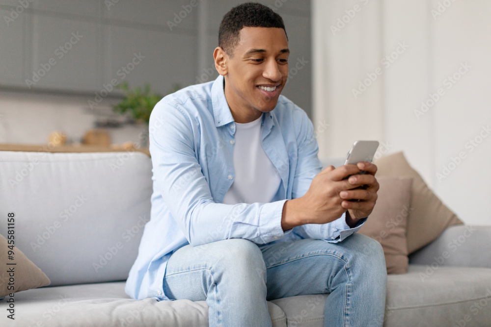 Smiling black guy using smartphone, browsing internet or texting, sitting on couch at home interior. Man using new cool application
