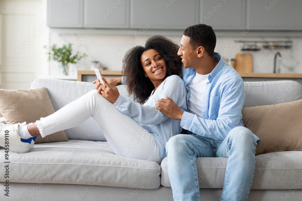 Happy black couple using smartphone, relaxing on couch at home, using new mobile application, browsing internet, checking social media Happy black couple using smartphone, relaxing on couch at home, using new mobile application, browsing internet, checking social media