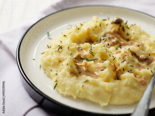 A plate of creamy mashed potatoes topped with a rich, savory gravy and garnished with fresh herbs. The dish is served on a white plate, with a spoon placed on the side, ready to be enjoyed