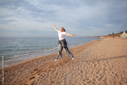A beautiful middle-aged woman walks on a sandy beach in autumn or spring. A 45-year-old woman is wearing jeans, a beige jacket and a plaid shirt. The active model jumps