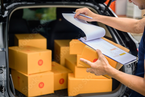 A delivery man stands at the back of a truck holding a clipboard and cardboard boxes, checking the address and cardboard boxes to confirm delivery