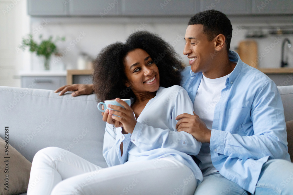 Happy African American spouses resting on sofa and talking, woman holding cup, enjoying relaxed weekend morning at home Happy African American spouses resting on sofa and talking, woman holding cup, enjoying relaxed weekend morning at home