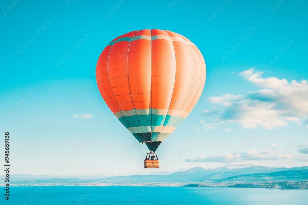 Vibrant orange and blue hot air balloon gracefully floating in a serene, clear blue sky over a vast, tranquil body of water with fluffy white clouds in the far distance. Vibrant orange and blue hot air balloon gracefully floating in a serene, clear blue sky over a vast, tranquil body of water with fluffy white clouds in the far distance.