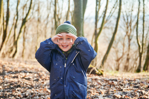 Young child smiling while hiking on forest trail, bundled up in blue winter jacket with fur-lined hood and green striped beanie. Kid enjoying sunny day outdoors.
