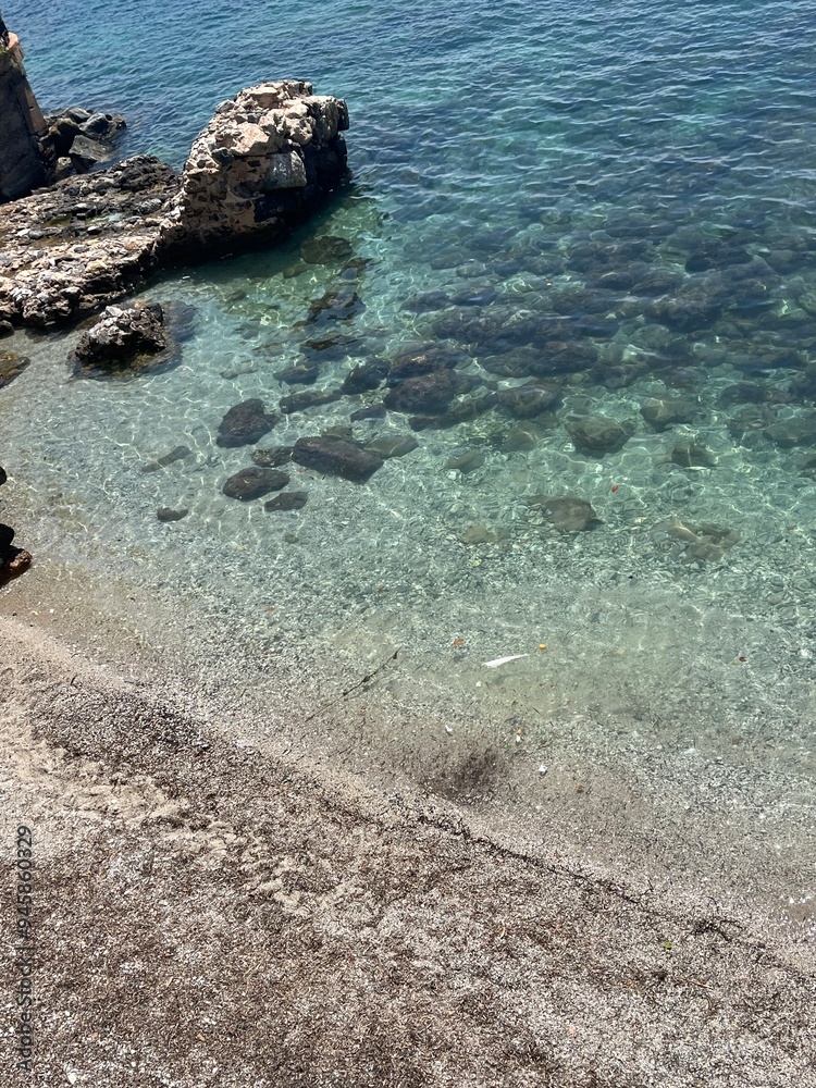 Obraz premium A serene shoreline with smooth pebbles and rocks meeting clear, turquoise water, highlighting the natural beauty of the coast. Perfect for a peaceful seaside escape.