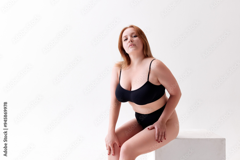 Fototapeta premium Portrait if attractive red-headed young woman with overweight body, sitting and confidently posing in underwear isolated on white studio background. Concept of female beauty, body positivity, care