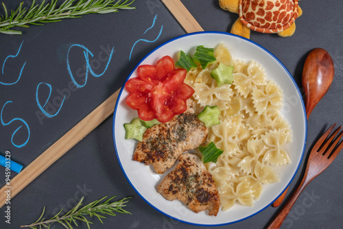 Plate of pasta, chicken chops, fresh vegetables for kid's lunch over black board background with inscription 