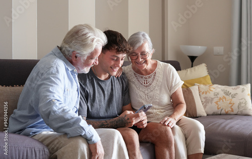 Happy Multi generation family of white haired grandparents and young grandson looking at something funny on nephew's mobile phone sitting together at home.