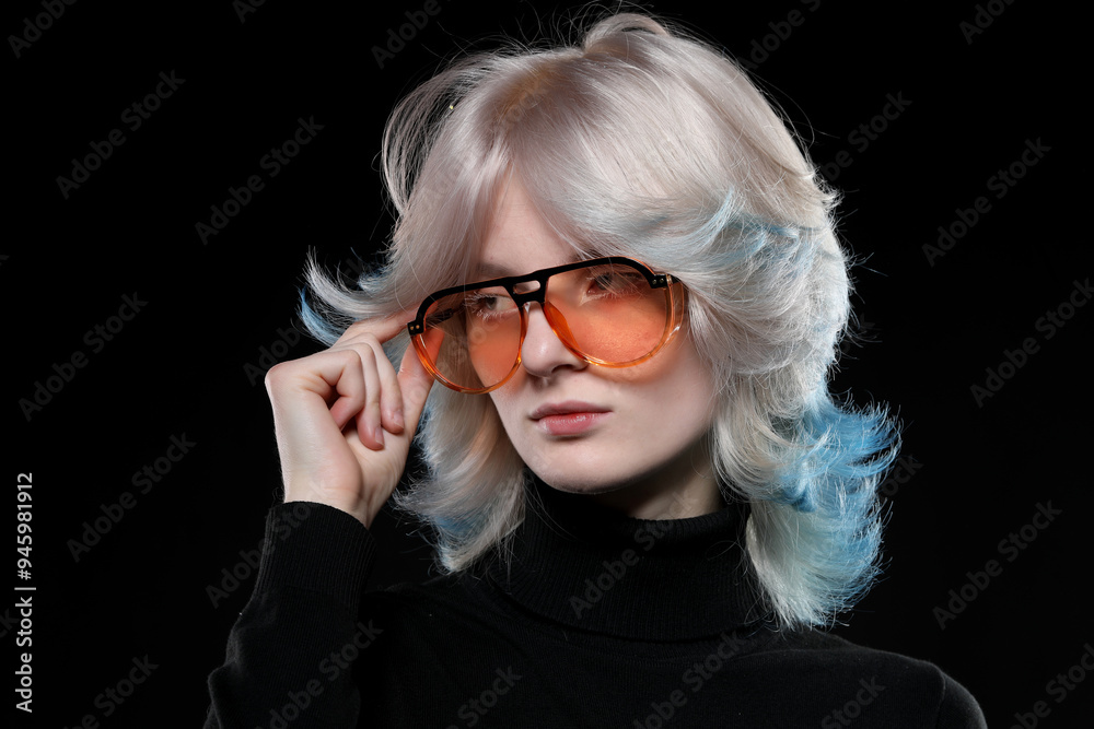 Beautiful young woman with creative hair coloring in blue streaks wears orange sunglasses, poses on black background. Beautiful young woman with creative hair coloring in blue streaks wears orange sunglasses, poses on black background.
