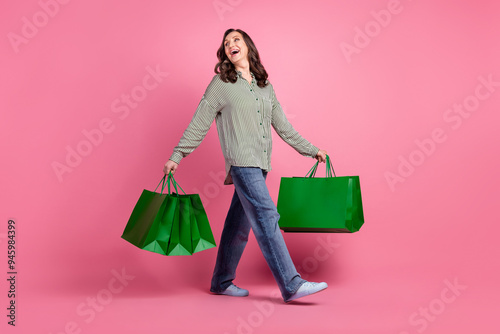 Full length photo of adorable carefree woman wear striped shirt holding shoppers walking empty space isolated pink color background