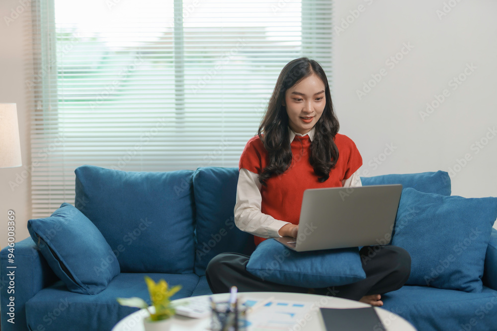 Young asian woman is working from home, sitting on a blue couch and using a laptop, with paperwork on the coffee table in front of her