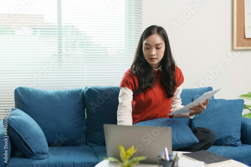 Young asian businesswoman is sitting on a blue sofa working from home using a laptop computer and analyzing financial chart documents