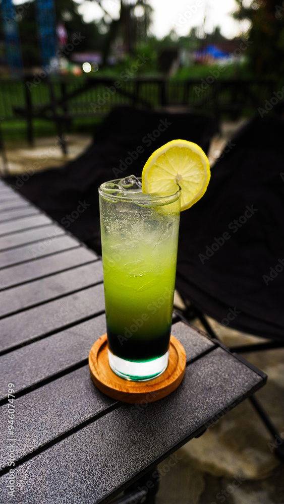 A refreshing green drink with a lemon wedge garnish sits on a wooden coaster on a table outdoors. A refreshing green drink with a lemon wedge garnish sits on a wooden coaster on a table outdoors.