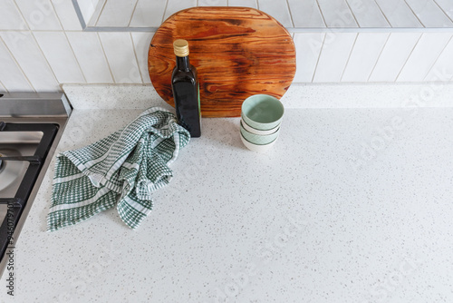 Styled surface on a modern white stone kitchen counter with white vertical wall tiles. Wooden cutting board, green kitchen towel cloth, small bowls and bottle of olive oil