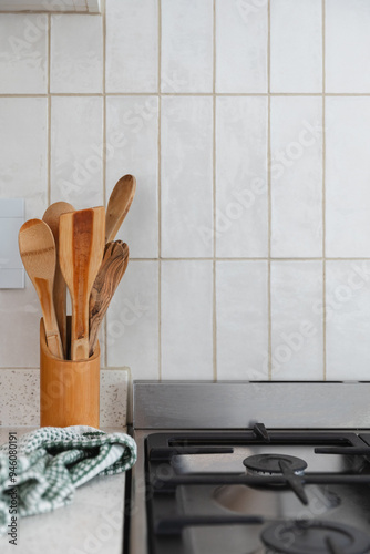 Wooden cooking utensils like spatula and spoon on a white stone kitchen surface next to a black and silver gas stove with white vertical wall tiles in the background