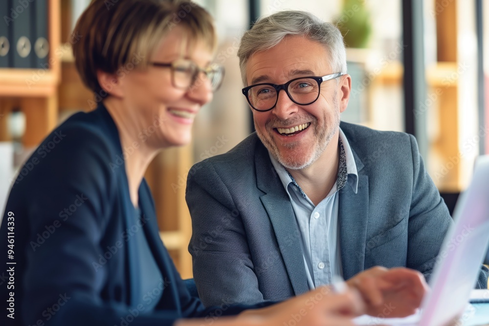 Two middle-aged colleagues smiling confidently at the office. Happy business partners working together, man and woman sharing a cup of hot drink, discussing project at the desk.