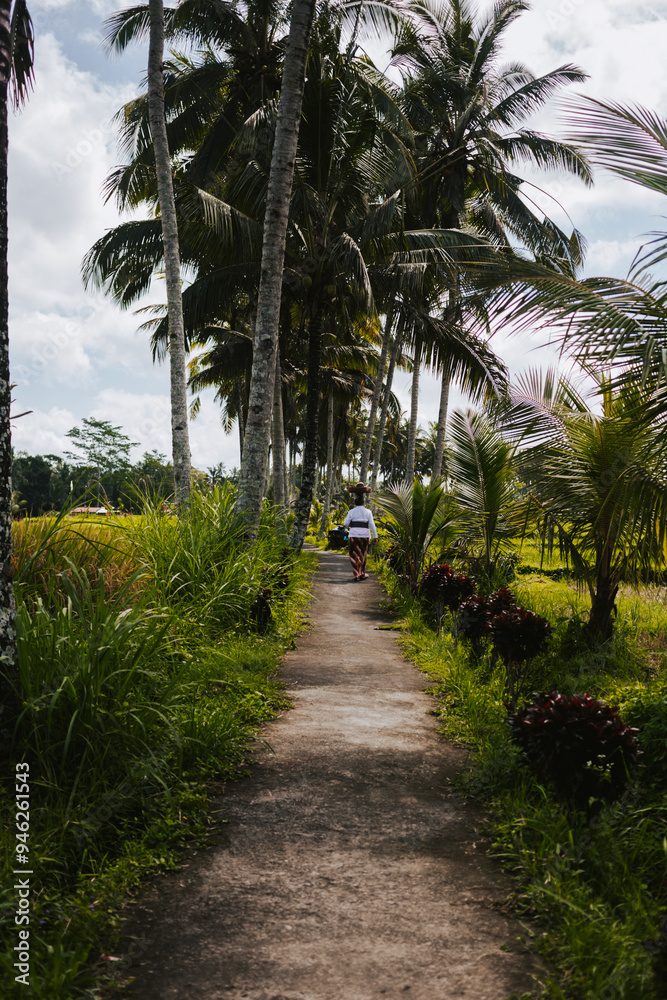 Obraz premium A woman in traditional Balinese clothes walks along the road against a backdrop of palm trees