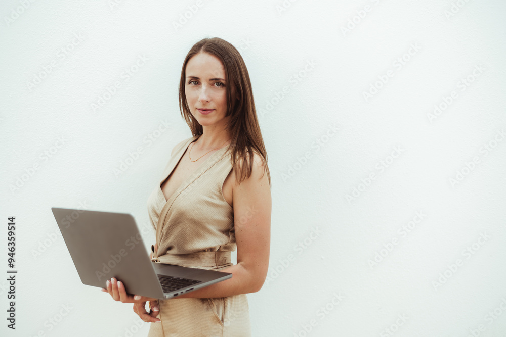 Portrait of a confident young hispanic woman holding a laptop against a plain light-colored background. She is wearing a beige dress, standing slightly angled, looking directly at the camera Portrait of a confident young hispanic woman holding a laptop against a plain light-colored background. She is wearing a beige dress, standing slightly angled, looking directly at the camera
