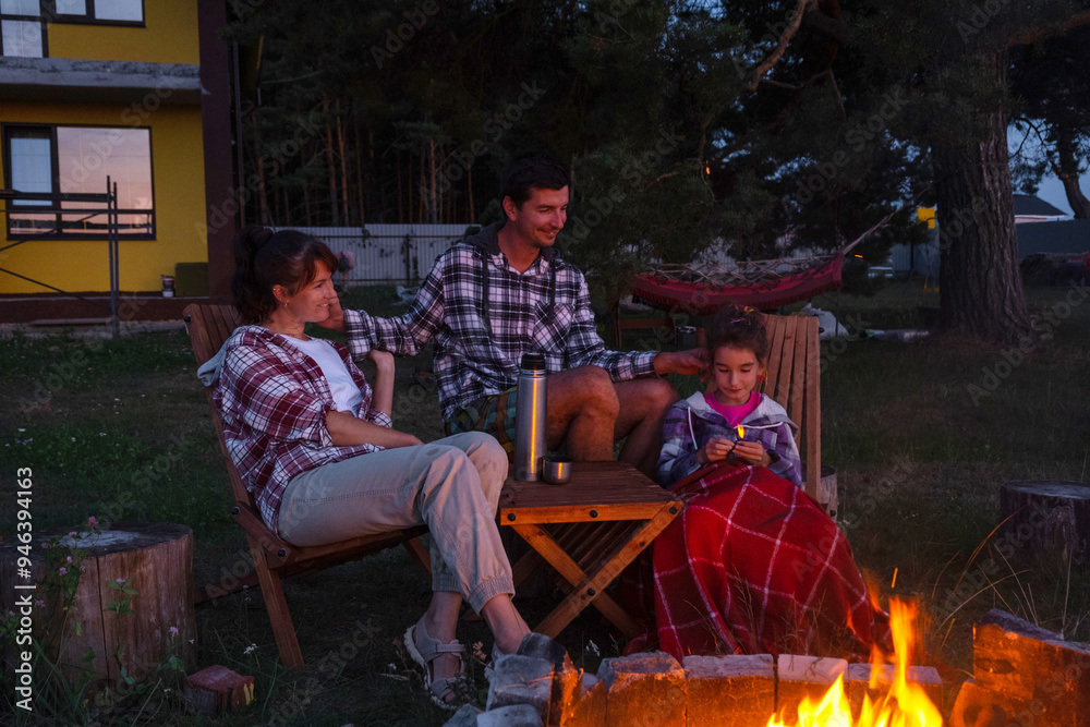 Naklejka premium Family is sitting by the outdoor campfire in the courtyard of the house on camping chairs, drink tea from a thermos, warm up and chat by the hearth