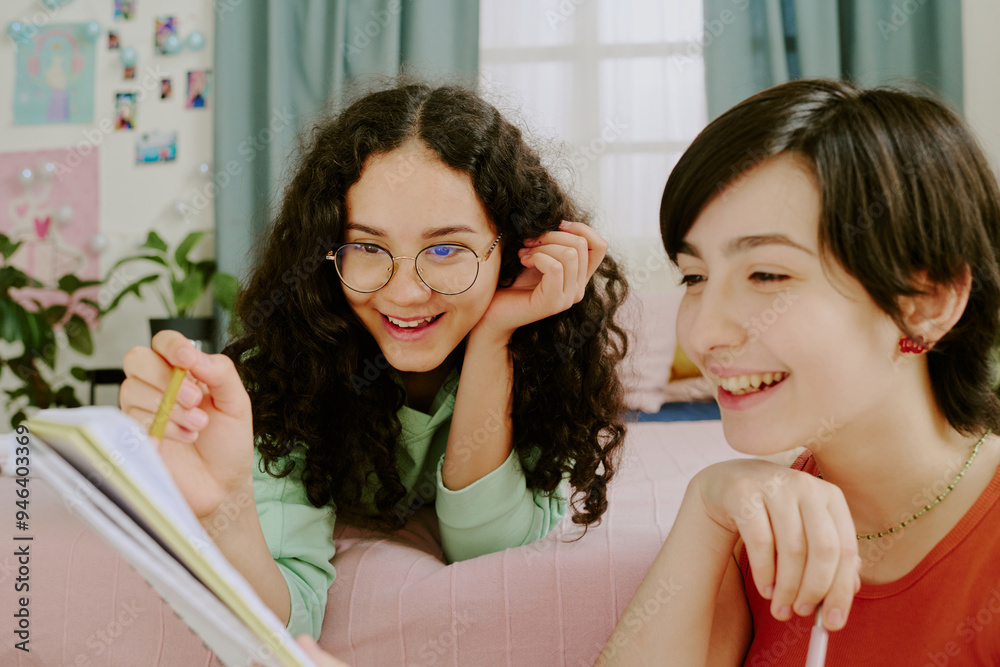Two young girls are sitting close, sharing a story and smiling warmly. Room is decorated with plants, photos, and colorful items, creating a cheerful ambiance Two young girls are sitting close, sharing a story and smiling warmly. Room is decorated with plants, photos, and colorful items, creating a cheerful ambiance