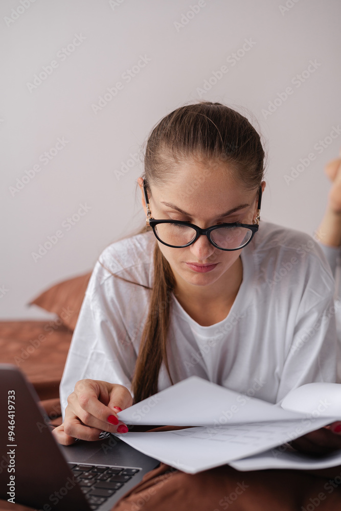 Young woman in glasses working remotely with documents and papers using laptop to check report at home Young woman in glasses working remotely with documents and papers using laptop to check report at home
