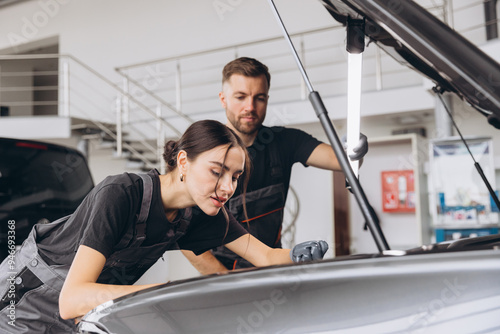 Trying to identify the source of the problem. Two mechanics man and woman working together at open hood on a car in an auto repair shop.