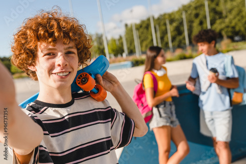 Smiling redhead curly teen boy in sporty casual outfit holding skate long board skateboard take selfie photo picture with his friends classmates in skate part urban street outside after school lessons photo