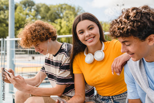 Group of teenagers students high school pupils classmates sitting outside yard using mobile app internet holding different gadgets smart phone hanging out in sport court park near campus photo