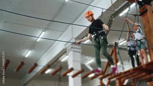 Wallpaper Mural A sporty 14-year-old girl with a smile on her face in protective gear moves along a rope bridge in a rope park indoors. A pretty girl in an orange helmet is engaged in extreme sports. High quality 4k Torontodigital.ca