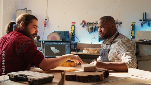 Happy BIPOC cabinetmaker and colleague comparing wood piece with schematic displayed on laptop. Cheerful carpenters in joinery crosschecking wooden object with notebook blueprint photo