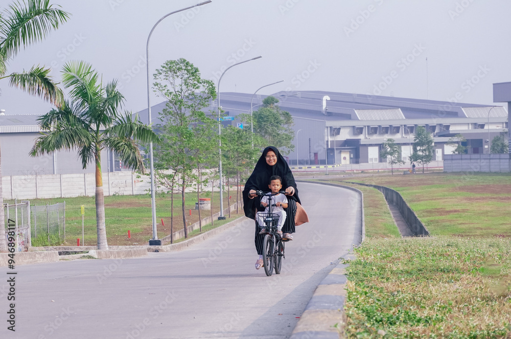 Asian mother in hijab riding a bicycle with her child on an urban road, showcasing family bonding and outdoor activities Asian mother in hijab riding a bicycle with her child on an urban road, showcasing family bonding and outdoor activities