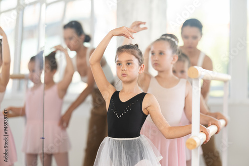 Adult woman teacher corrects group of girls dancers doing batman tandu movement at ballet barre photo