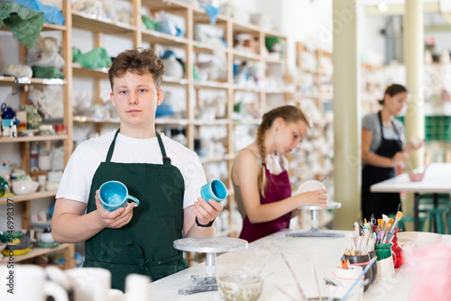 Guy teenager student in apron posing with ceramic cups in his hands in ceramic workshop