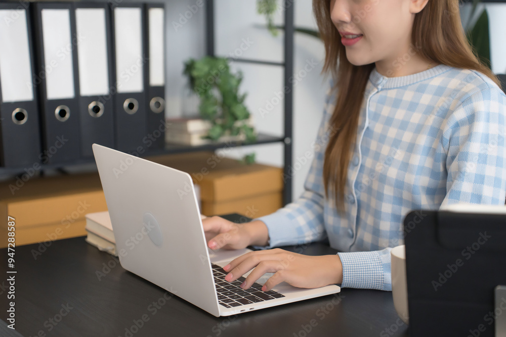 An Asian female businesswoman in a blue-checkered shirt is focused on working on her laptop at a modern office desk, surrounded by neatly organized files and office supplies An Asian female businesswoman in a blue-checkered shirt is focused on working on her laptop at a modern office desk, surrounded by neatly organized files and office supplies