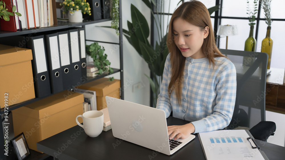 An Asian female businesswoman in a blue-checkered shirt is focused on working on her laptop at a modern office desk, surrounded by neatly organized files and office supplies An Asian female businesswoman in a blue-checkered shirt is focused on working on her laptop at a modern office desk, surrounded by neatly organized files and office supplies