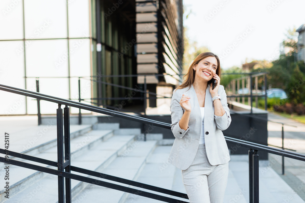 Fototapeta premium Woman enjoys a sunny outdoor stroll while engaged in a lively phone conversation, exuding confidence in a stylish gray outfit