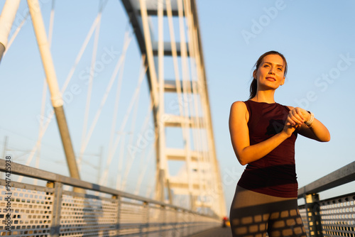 Female runner checks her watch while standing on a sunlit bridge during the golden hour, reflecting her dedication to fitness and training outdoors