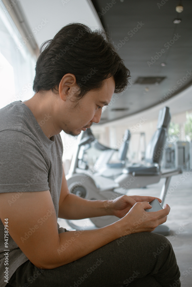 Fototapeta premium Vertical photo of asian Thai man sitting at gym and using mobile phone after done or between resting exercise, workout with dumbbells.