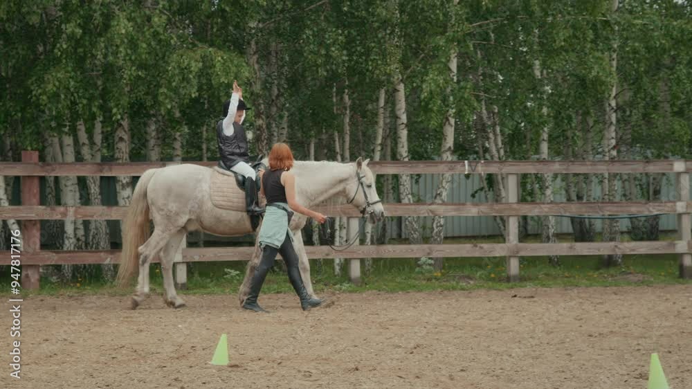 Full shot of child with Down syndrome wearing equestrian attire sitting astride white mare while having horse riding lesson with young female coach on outdoor riding arena