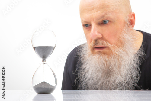 A contemplative man with a long white beard intently observes an hourglass, reflecting on the passage of time. The minimalist white background highlights the stark contrast between  photo