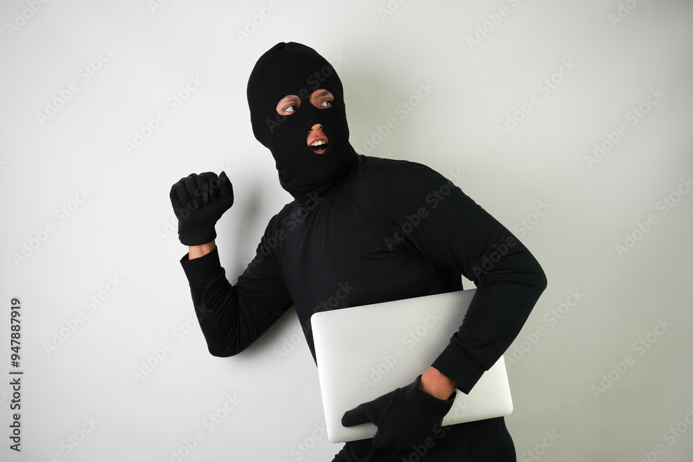 Young Adult black masked robber is holding silver colored laptop. Running, fleeing and escaping pose. Concept for hacking, scamming, phishing. Young Adult black masked robber is holding silver colored laptop. Running, fleeing and escaping pose. Concept for hacking, scamming, phishing.