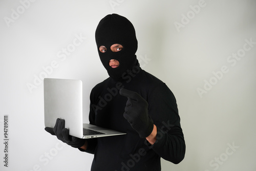 Young Adult black masked robber wearing gloves is holding silver colored laptop for hacking, scamming and phishing.