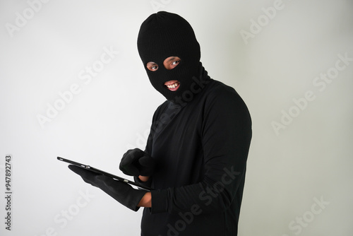 Young Adult black masked robber is holding digital tablet with blank green screen and he calling his target victim with terror. Concept for hacking, scamming, phishing.