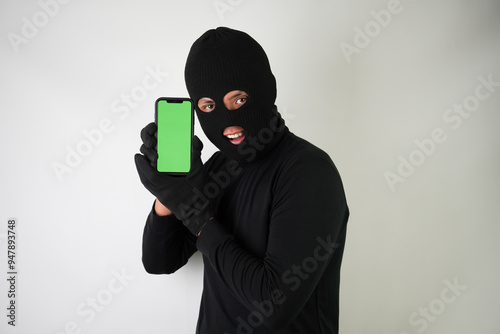 Young Adult black masked robber is holding smartphone with blank green screen. Concept for hacking, scamming, phishing.