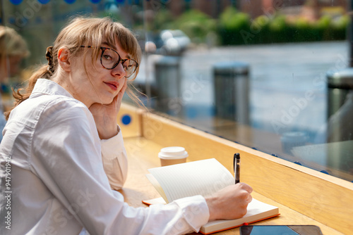 Young Woman in Glasses Thoughtfully Journaling at Cozy Café on Sunny Afternoon photo