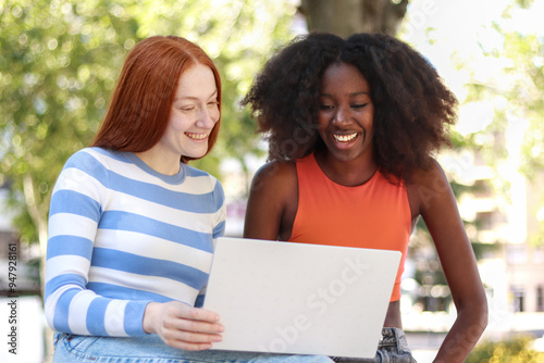 Multiracial two women friends laughing together using a laptop in a sunny park