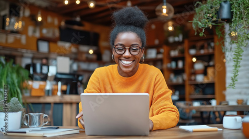 A woman wearing glasses and a bright orange sweater is smiling and working on a laptop in a cozy cafe. Various plants and warm lighting create a comfortable atmosphere.